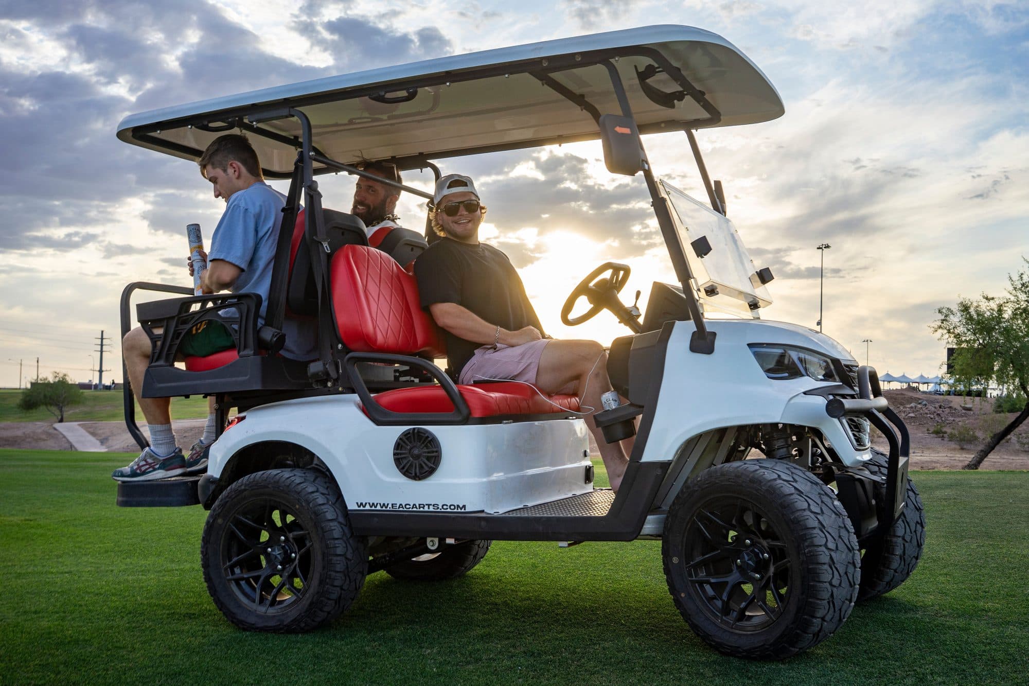White EA4R+ 60V four-seat lifted lithium electric golf cart with red seats and black lifted wheels carrying three adult passengers on a golf course at sunset
