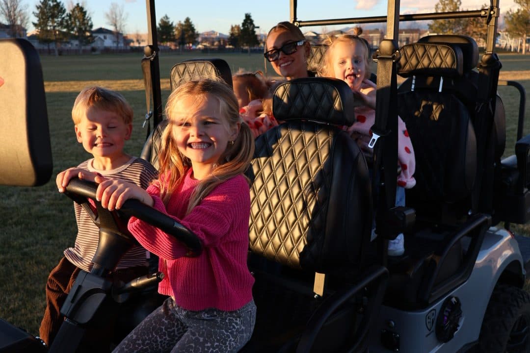 Children smiling at the wheel of a gray EA Carts golf cart with black quilted seats, an adult behind them