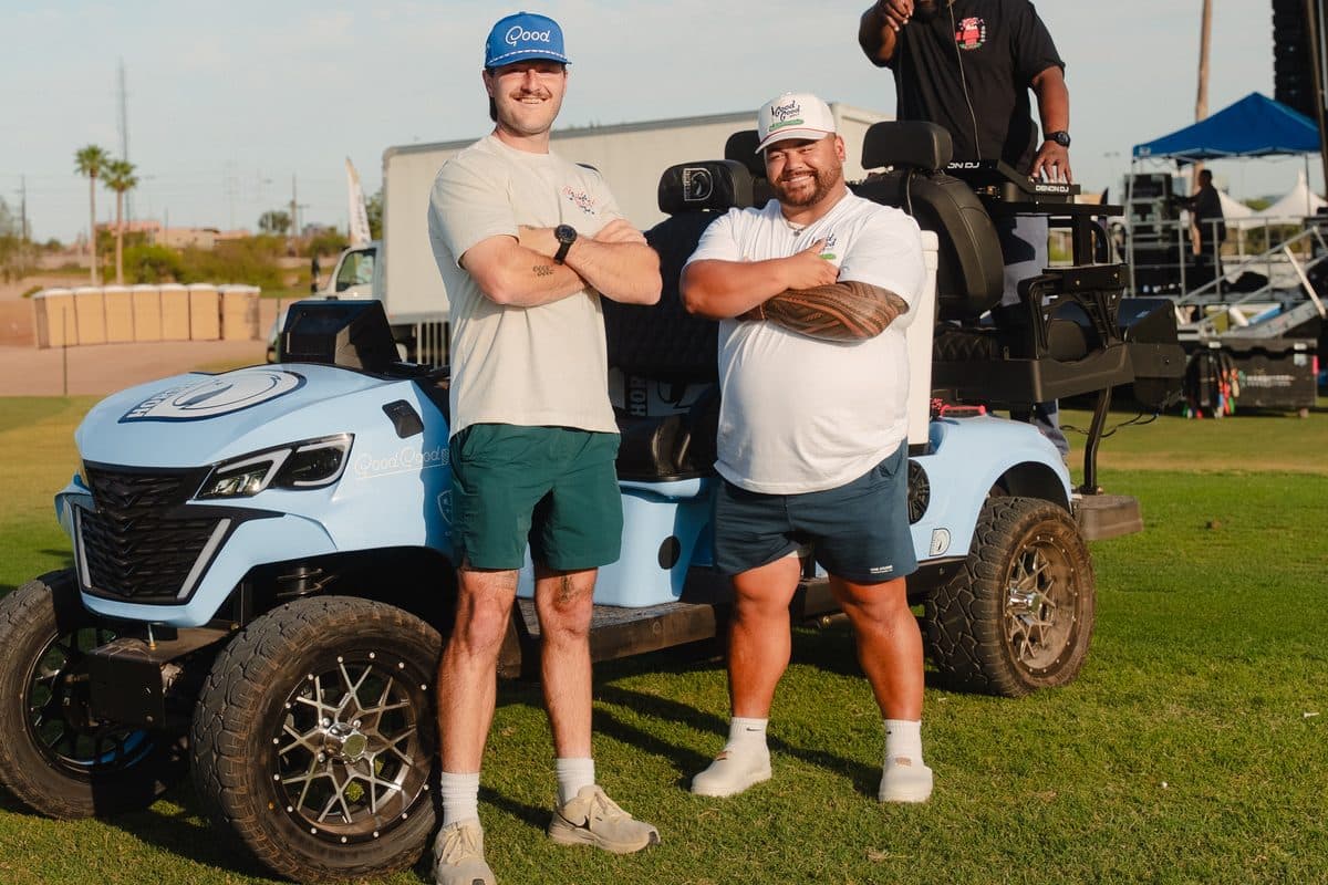 Two adults with arms crossed in front of a light blue Good Good custom EA Carts golf cart on grass