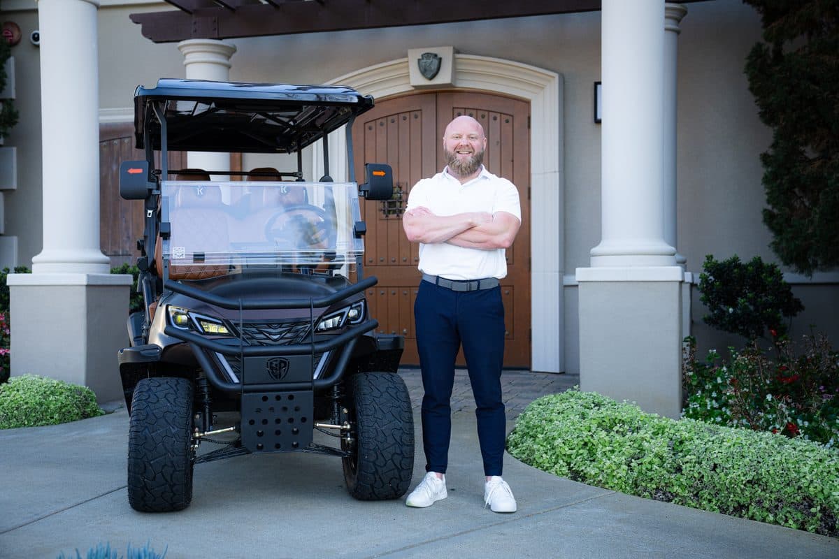 TJ Johnson, arms crossed, beside a black EA Carts build with tan quilted seats at a building entrance with wooden doors