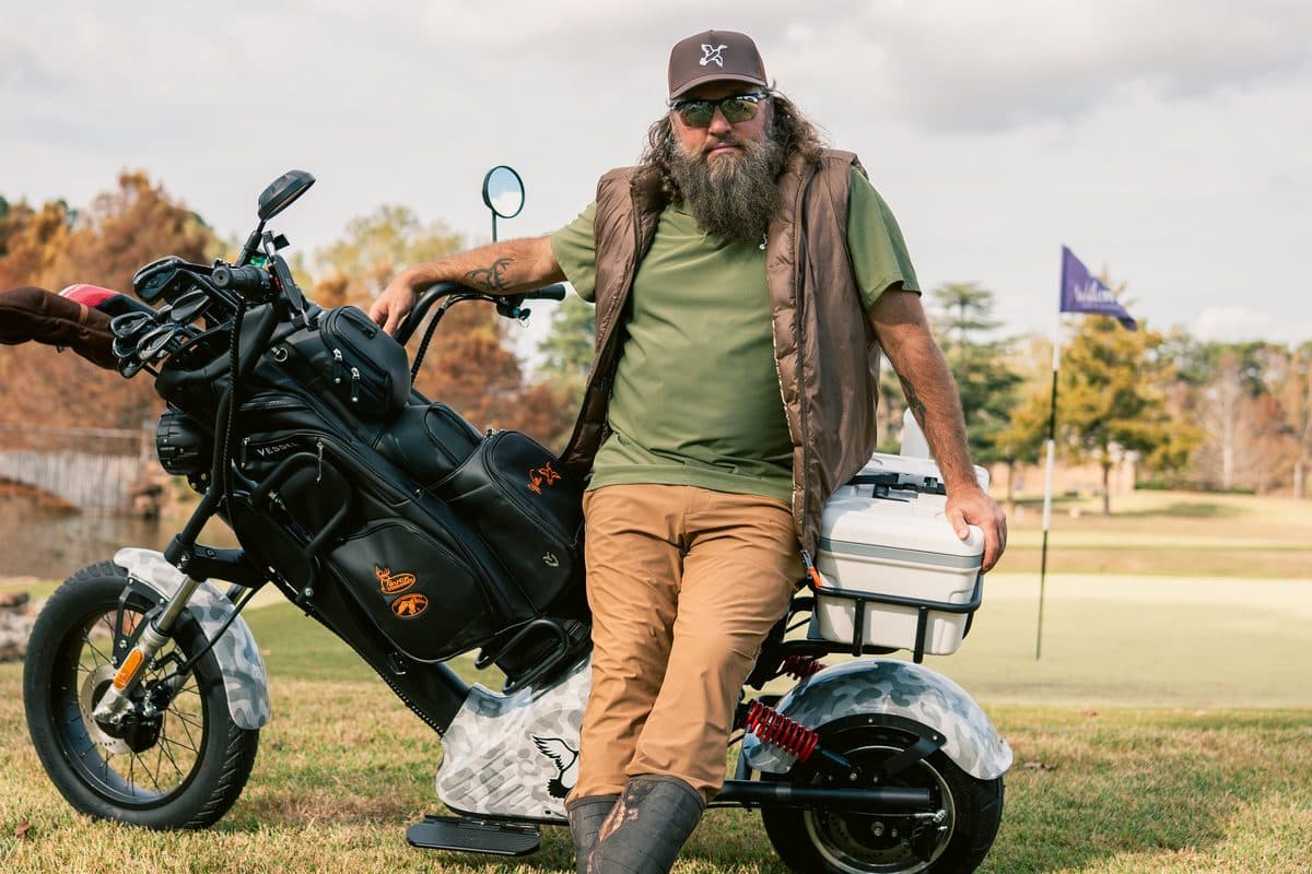Bearded adult in a cap and vest leaning on a duck-camo wrapped EA Golf Bike with a black golf bag on a golf course