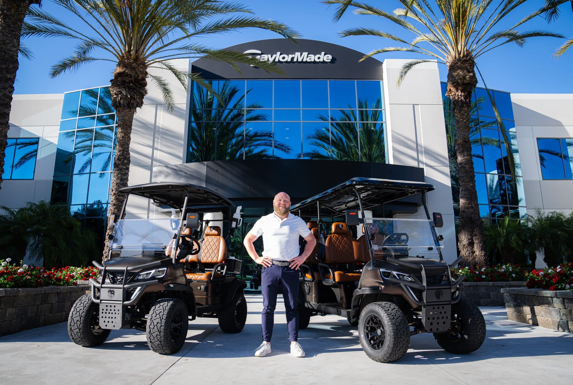 TJ Johnson standing between two black EA Carts builds with tan quilted seats in front of the TaylorMade headquarters sign